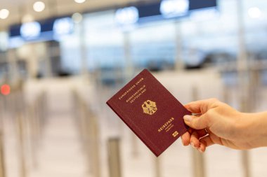 Bir kişi kontrol gişeleriyle dolu bir havaalanı terminalinde dururken elinde pasaport tutuyor. Gezginlerin yaklaşan uçuşlarına hazırlandıkları görülebilir..