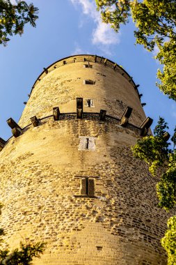 Güçlü bir taş kule, yemyeşil ağaçlarla çevrili gökyüzüne doğru uzanır. Güneş ışığı, kulelerin yıpranmış dokusunu vurgular. Derin mavi gökyüzü ise dingin bir zemin sağlar..
