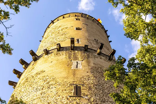 Uzun taş kule gururla yükseliyor açık mavi gökyüzüne ve canlı yeşil ağaçlara karşı, yıpranmış yapısını ve dalgalanan bayrağını sergiliyor..