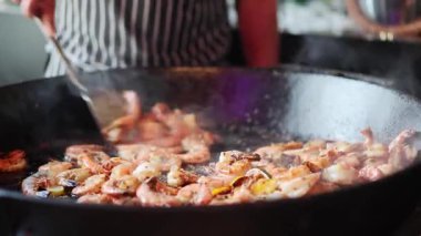 Bir şef geniş bir mutfakta karidesi ustalıkla hazırlar ve büyük bir tavada karıştırır. Karides cızırdarken buhar yükselir ve lezzetli bir atmosfer yaratır..