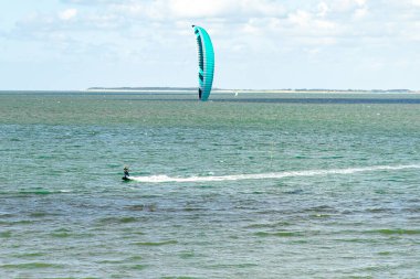 Yanlız bir uçurtmacı, büyük mavi bir uçurtma tarafından itilen, ışıldayan suyun üzerinde kayar. Parlak gökyüzü ve nazik dalgalar bu heyecan verici etkinlik için mükemmel bir yaz atmosferi yaratıyor..