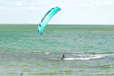 Uçurtma sörfçüsü ustalıkla havada süzülen canlı bir uçurtmayla ışıldayan turkuaz suyun üzerinde manevra yapar. Sahne güneşli bir öğleden sonrayı yakındaki heyecanla yakalıyor..