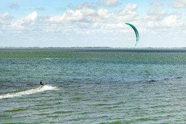 Yetenekli bir uçurtma sörfçüsü güneşli bir günde sakin sularda manevra yapar. Ufuk, üzerinde yumuşak dalgalar ve kabarık bulutlar bulunan bir kıyı zeminine sahiptir..