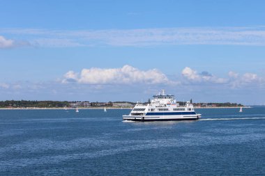 Feribot, uzak bir kıyı şeridi ile sakin sularda sorunsuzca hareket eder. Yelkenliler, parlak mavi gökyüzünün altında tüylü bulutlarla çevrili bir atmosfer yaratıyorlar..
