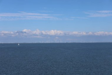 Küçük bir tekne berrak mavi bir gökyüzünün altında sakin sularda süzülüyor. Rüzgâr türbinleri uzak ufuktan görülebiliyor. Huzurlu manzaraya katkıda bulunuyorlar..