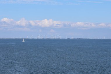 Yalnız bir yelkenli sakin sularda zarifçe hareket eder. Ufukta parlak, parçalı bulutlu gökyüzünün altında bir dizi rüzgar türbini görünür..