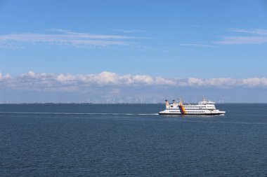 Bir feribot sakin sularda sorunsuzca yol alır. Ufukta, dağınık bulutlarla dolu parlak mavi bir gökyüzünün altında rüzgâr türbinleri görünür. Huzurlu bir anı yakalar.