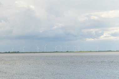 Açıklardaki rüzgar türbinleri kıyı şeridi boyunca stratejik olarak sakin sularla çevrili ve karamsar, bulutlu bir gökyüzü ile çevrili. Bu yenilenebilir enerjiye olan bağlılığı vurgular.