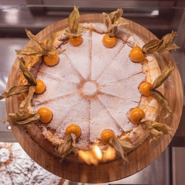 A beautifully crafted cake sits on a wooden platter, adorned with fresh fruit and powdered sugar. The cozy bakery ambiance highlights this tempting dessert during the afternoon.