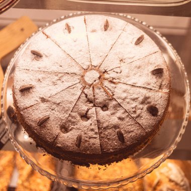 Güzel yuvarlak bir pasta, pudra şekerle tozlanmış ve bademlerle süslenmiş bir pastanede cam altında oturur. Tatlı cazibesi ve davetkar sunumuyla ziyaretçileri cezbediyor..
