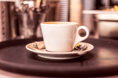 Taze kahveyle dolu beyaz bir kahve fincanı yanında kaşıkla küçük bir tabakta oturur. Ortam sıcak ve davetkar, sabah molası için mükemmel..