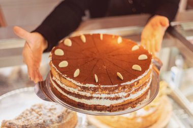 A beautifully crafted layered cake with white cream and almond decorations is served by a bakery staff member in a cozy atmosphere during the afternoon.