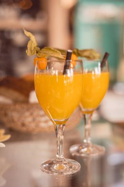 Meyve ve yapraklarla süslenmiş iki canlı portakal kokteyli, müşteriler hareketli bir kafede güneşli bir öğleden sonranın tadını çıkarırken yansıtıcı bir yüzeyde dinleniyor, ferahlık ve neşe anlarını kutluyorlar..
