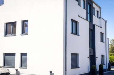 A modern white building stands in a quiet neighborhood. It features large windows and clean lines, showcasing a contemporary architectural style in bright daylight.