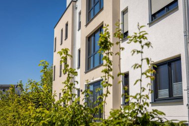 Parlak güneş ışığı, temiz hatları ve geniş pencereleriyle modern bir apartmanı aydınlatıyor. Yeşil çalılar sahneyi çerçeveler, davetkar atmosferi güçlendirir.