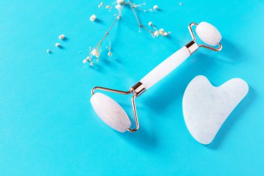 Mavi arka planda çingene çiçekleri olan gül kuvars yüz masajı silindiri. Yeşim taşlı masaj aletleri, kırışıklık önleyici cilt bakımı aletleri. Boşluğu kopyala.