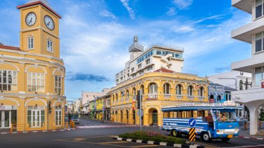 Phuket, Tayland - 11 Kasım 2024: Phuket Eski Kasabası 'ndaki ikonik sarı saat kulesi ve tarihi Chartered Bank binası, Sino-Portekiz mimarisini sergiliyor, oradan geçen mavi bir yerel otobüsle hareketli bir kavşak çerçevesi oluşturuyor.