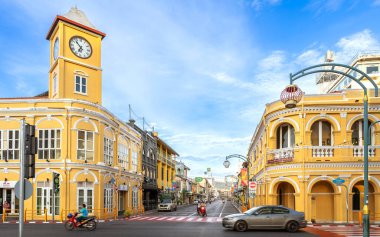 Phuket, Tayland - 11 Kasım 2024: Phuket Eski Kasabası 'nda Sino-Portekiz mimarisinin yer aldığı ikonik sarı saat kulesi, açık mavi gökyüzü altında hareketli bir cadde kavşağının merkezinde duruyor.