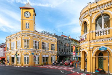 Phuket, Tayland - 11 Kasım 2024: Phuket Eski Kasabası 'ndaki Sino-Portekiz mimarisinin ayırt edici özelliği olan ikonik sarı saat kulesi, araçlar ve motosikletlerle dolu hareketli bir kavşağın yanında duruyor.