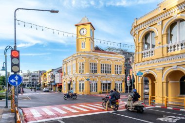 Phuket, Tayland - 11 Kasım 2024: Phuket Eski Kasabası 'ndaki ikonik sarı saat kulesi, Çin-Portekiz mimarisini gösteriyor, motosikletler ve yayalarla dolu bir kavşağın yanında duruyor.