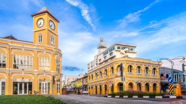 Phuket, Tayland-10 Kasım 2024: Phuket Eski Kasabası 'ndaki tarihi Sino-Portekiz saat kulesi ve Kiralık Banka binası açık mavi gökyüzünün altından geçen kendine özgü mavi bir yerel otobüsle hareketli bir kavşak oluşturuyor.