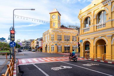 Phuket, Tayland - 10 Kasım 2024: Phuket Eski Kasabası 'ndaki ikonik sarı saat kulesi, Çin-Portekiz mimarisini gösteriyor, motosikletler ve yayalarla dolu bir kavşağın yanında duruyor.