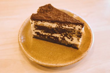 Yumuşak odaklı lezzetli tiramisu keki sıcak ve rahat bir kafe atmosferindeki ahşap bir masaya konmuş seramik bir tabağa servis edilir..