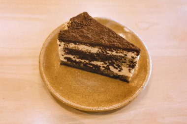 Yumuşak odaklı lezzetli tiramisu keki sıcak ve rahat bir kafe atmosferindeki ahşap bir masaya konmuş seramik bir tabağa servis edilir..