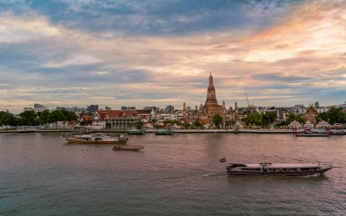 Bangkok, Tayland-5 Temmuz 2025 Chao Phraya Nehri 'nin karşısındaki Wat Arun Tapınağı' nın dramatik bir günbatımı gökyüzünün altından geçen tekneleri.