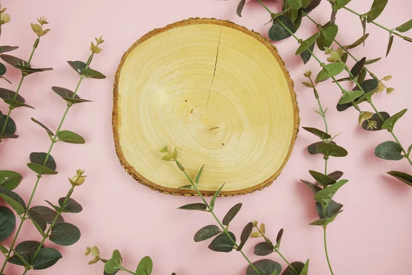 Ahşap podyum ürün görüntüsü düz duruyor. Pembe arkaplanda yapraklar var.