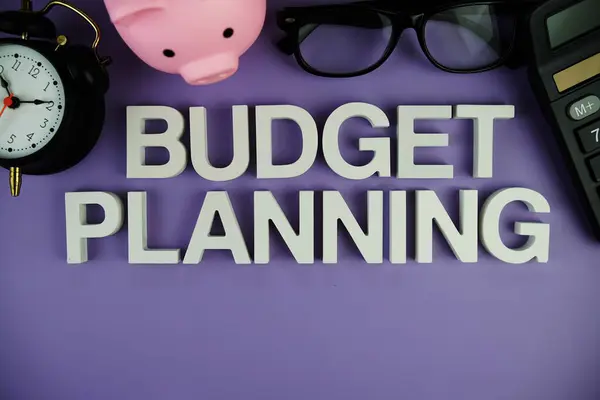 Budget Planning alphabet letters with piggy bank, alarm clock, calculator top view on purple background