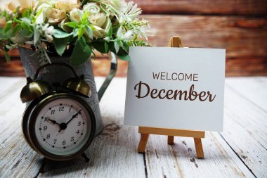 Hoş geldiniz, aralık ayında kağıt kartın üzerine ahşap sehpa ve metal vazo süslemesi çiçekli alarm saati ile yazılmış mesaj.