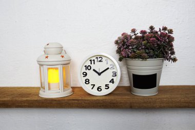 Beyaz fener, beyaz alarm saati ve ahşap raflarda yapay bitki.