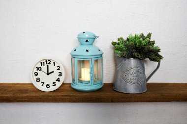 Mavi fenerli beyaz saat ve tahta raflarda yapay bitki.