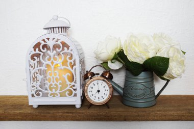 Ev dekorasyonu aksesuarları LED mum ışığı, çalar saat ve ahşap raflardaki metal vazodaki çiçek ve beyaz beton duvarlı beyaz fener.