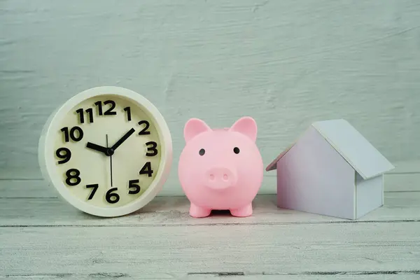 Piggy bank with house model and white clock on wooden backgroun, business background