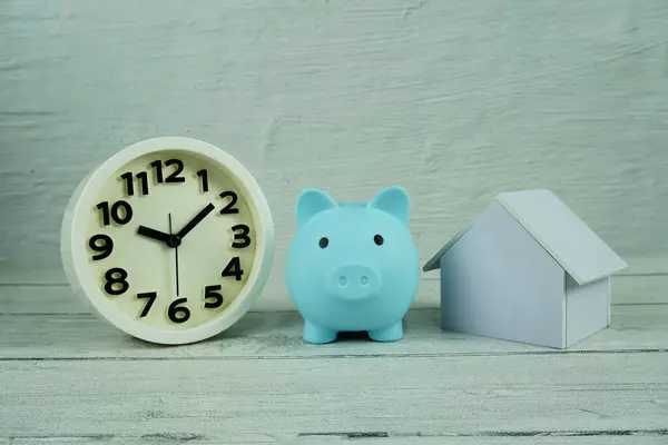 Piggy bank with house model and white clock on wooden backgroun, business background