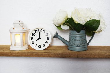 Ev dekorasyonu aksesuarları beyaz fener, çalar saat, ahşap raflardaki metal vazodaki çiçek ve beyaz beton duvar.