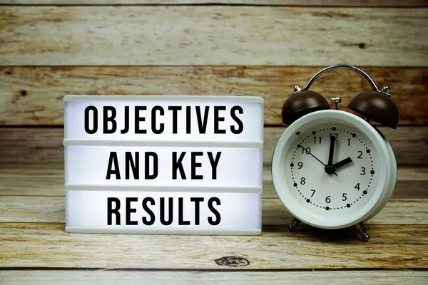 Stock image OKR - Objective Key Results text on LED lightbox with alarm clock on wooden background