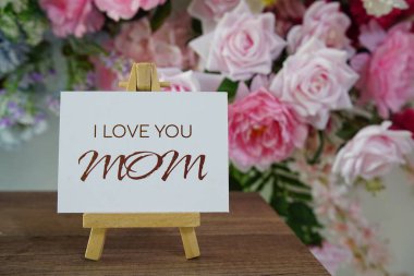 Seni seviyorum, Anneler Günün kutlu olsun. Kağıt karta tahta sehpa ve ahşap arka planda çiçek süslemesi yazılı.