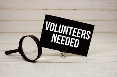 Volunteers Needed text message on paper card with magnifying glass on wooden background clipart