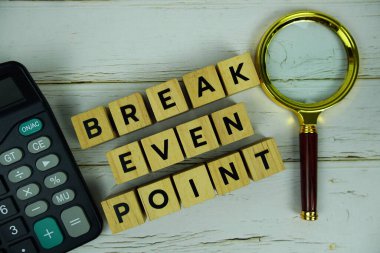 Break Even Point with wooden blocks alphabet letters and magnifying glass, calculator top view on wooden background clipart