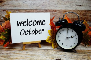 Alarm saati ve ahşap arka planda akçaağaç yaprağı süslemesiyle Ekim ayına hoş geldiniz metni