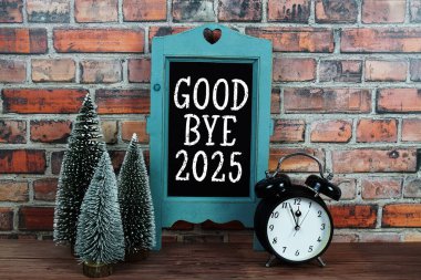 Elveda karatahta sehpadaki 2025 metni. Alarm saati ahşap ve eski tuğla duvar arkaplanlı.