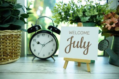 Pencerenin arka planında resim sehpası ve alarm saati olan kağıt kart üzerindeki Temmuz ayına hoşgeldiniz