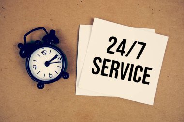 Kağıt işçiliği arka planında alarm saati olan yapışkan notla 24 / 7 servis metni