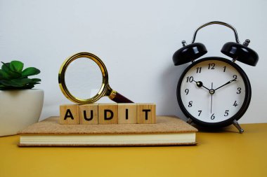 Tahta bloklar alfabe harfleriyle büyüteç ve alarm saati sarı arkaplanda AUDIT metin mesajı