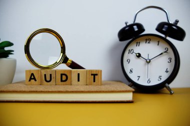 Tahta bloklar alfabe harfleriyle büyüteç ve alarm saati sarı arkaplanda AUDIT metin mesajı