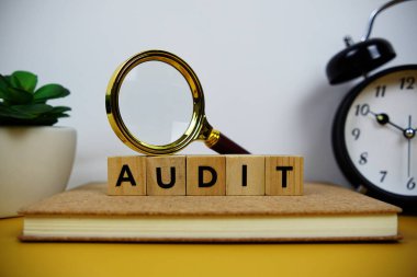 Tahta bloklar alfabe harfleriyle büyüteç ve alarm saati sarı arkaplanda AUDIT metin mesajı