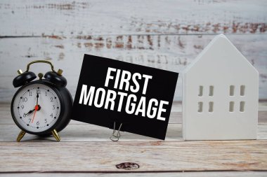 Kağıt kartın üzerindeki ilk ipotek, alarm saati ve ahşap arka planda beyaz ev modeli.